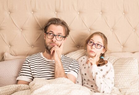 Thoughtful or bored caucasian father and daughter wearing pyjamas and glasses in a bed. Morning in a bedroom - family time.の写真素材
