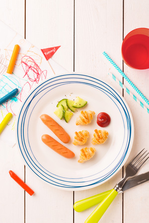 Kid's breakfast - weenies, mini croissants and cucumber salad. Plate captured from above (top view, flat lay) on white wooden table. Layout with free copy (text) space.の写真素材