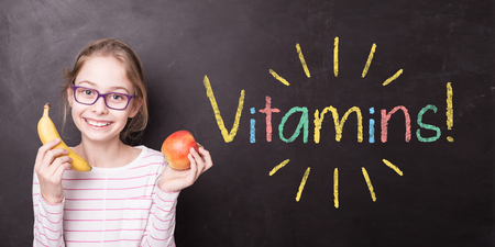 Happy smiling blond child girl holding fruits (apple and banana). School chalkboard with 'Vitamins!' sign as background. Healthy snack - diet, nutrition and eating habits - motivational poster design.の写真素材