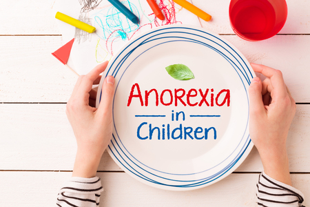 Plate with a leaf and 'Anorexia in Children' sign in kid's hands. Eating disorders - concept captured from above (top view, flat lay). Crayons and child's drawing around.の写真素材