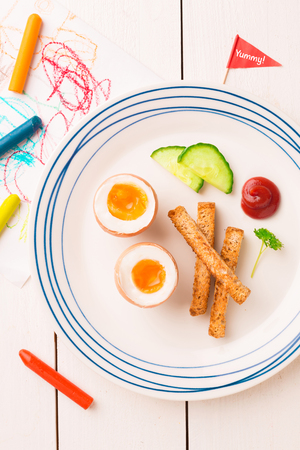 Kid's breakfast - eggs, toasts, cucumber and ketchup. Plate captured from above (top view, flat lay) on white wooden table.の写真素材