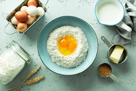 Kitchen - preparing dough. Bowl with flour and eggs. Basic food cooking ingredients around (butter, milk, sugar) on pastel blue stone background. Captured from above (top view, flat lay).の写真素材