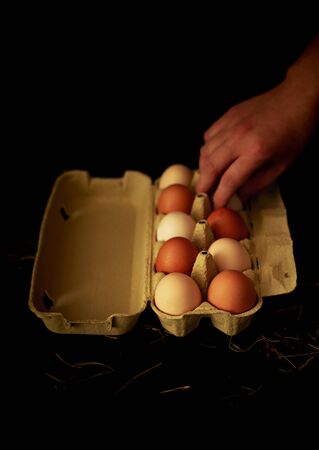 Food - hand reaches for an eggs in a carton box on black. Dark moody composition. Background with free copy (text) space.の写真素材