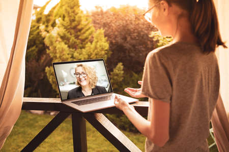 Caucasian teenage girl having video chat on laptop computer with her working mother while standing outdoor on the garden terrace at home. Technology - modern parenting and family lifestyle.の写真素材