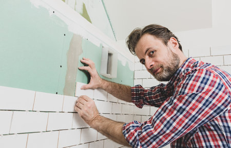 Caucasian interior finishing worker (tiler) laying small white ceramic tiles on the wall. Bathroom renovation works. Handyman in plaid shirt.の写真素材