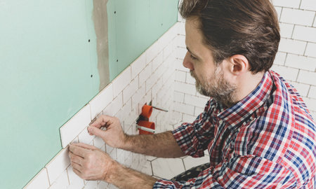 Caucasian interior finishing worker (tiler) laying small white ceramic tiles on the wall. Bathroom renovation works. Handyman in plaid shirt.の写真素材