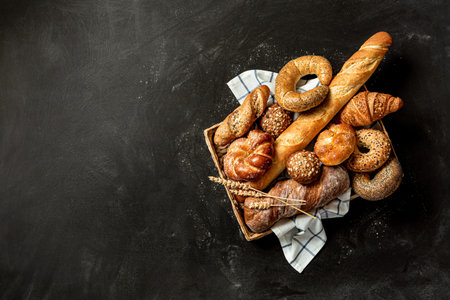 Bakery - various kinds of breadstuff on the tray. Bread rolls, baguette, bagel, sweet bun and croissant captured from above (top view, flat lay). Black chalkboard background with free copy space.の写真素材
