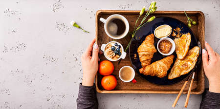Woman serving continental breakfast on wooden tray. Coffee, croissants, jam, butter, fruits and flowers captured from above (top view, flat lay). Stone background with free copy space.の写真素材