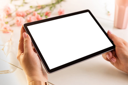 Woman's hands holding tablet with blank (empty) screen close up. Spring pastel pink flowers and...