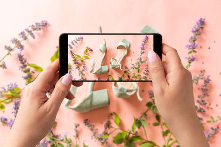 Woman taking photo of pastel mint green sandals shoes and meadow flowers with smartphone. Blogger, influencer or stylist capturing fashion footwear for social media. Pink background.の写真素材