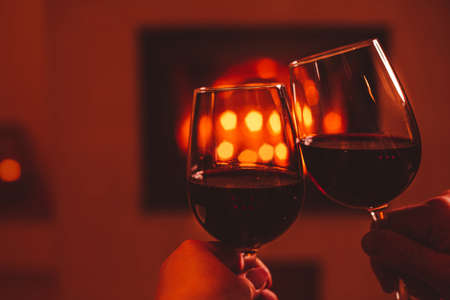 Romantic date - couple making a toast with two glasses of red wine. Fireplace as background - free copy (text) space. Christmas or Valentine's Day mood.の写真素材
