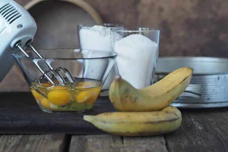 Stage of cooking a banana biscuit. Beat eggs with a mixer in a glass bowl. Ingredients for making biscuit on a brown ancient wooden background. Pastry backgroundの写真素材