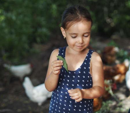 Little girl holding a leaf of a tree on a background of green trees in the countryside. Summer day.の写真素材
