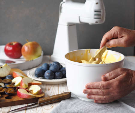 The process of making a charlotte with apples and plums. Hands of an elderly woman gently knead the dough with flour. Preparation for the holiday or for meeting guests.の写真素材