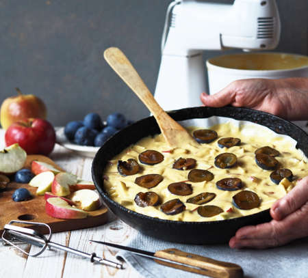 Elderly woman's hands hold a frying pan with apple and plum charlotte dough. Woman's hobby. Preparation for a holiday or meeting guests.の写真素材