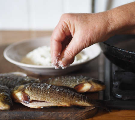 The process of preparing river fish crucian carp for frying in a frying pan. An elderly woman's hand salts the fish.の写真素材
