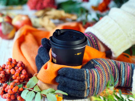 Hands in gloves hold a black glass of coffee on a background of autumn leaves.の写真素材