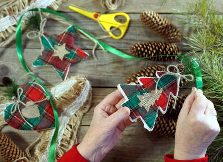 Female elderly hands are busy making New Year's toys. New Year's composition on a wooden background with pine branches, cones, handmade New Year's toys. Pine branches, rope, toys and scissors. New Year greetings background.の写真素材
