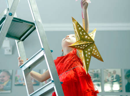 A little girl in a festive red dress is busy decorating the room with a paper gold star. New Year is eco friendly paper decor.の写真素材