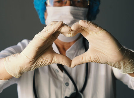 Kind elderly doctor in protective medical mask showing heart sign with hands in sterile gloves. The...