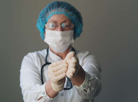 Studio photo of an elderly woman doctor wearing a mask in uniform putting on gloves against a gray background. Profession saving lives. Helping elderly doctors during the coronavirus pandemic.の写真素材