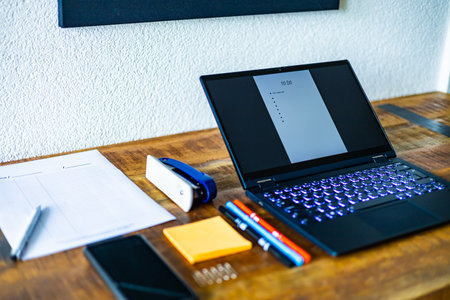 A laptop is open on a desk with a stack of papers and a pen. The laptop screen is displaying a list of tasksの写真素材