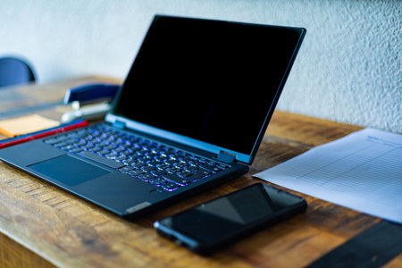 A laptop is open on a wooden desk with a cell phone and a notepad. The laptop is black and has a...