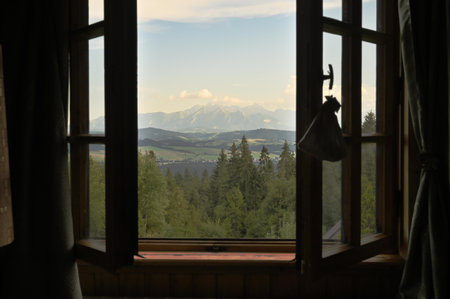 Beautiful view on green trees forest, blue sky and mountains through a wooden window frame of an old...