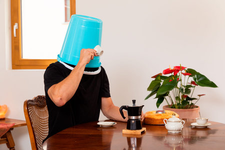 Man drinks coffee with a blue bucket on his head, flowers in the background and the coffee maker on the tableの写真素材