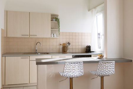 Kitchen with island with two white stools and a gray worktop. Bright window with curtain. Nobody...