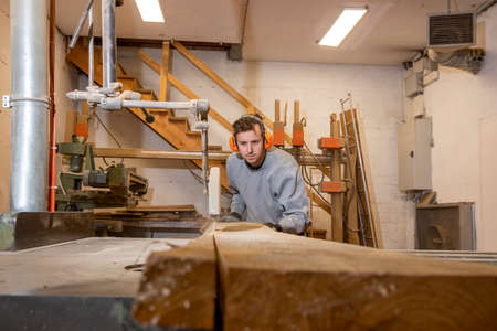Young and handsome carpenter boy, concentrated on his job while cutting a long plank of wood with a circular saw.の写真素材