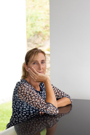 Portrait of a middle-aged woman with blond hair in a ponytail with a window with a green lawn and a white wall behind her. Reflective table and white backgroundの写真素材