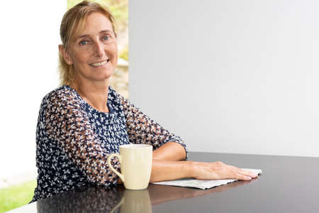 Portrait of a middle-aged woman with blond hair in a ponytail with a window with a green lawn and a white wall behind her. Reflective table and white backgroundの写真素材