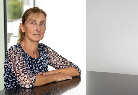 Portrait of a middle-aged woman with blond hair in a ponytail with a window with a green lawn and a white wall behind her. Reflective table and white backgroundの写真素材