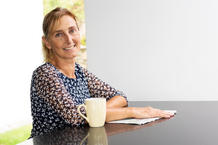 Portrait of a middle-aged woman with blond hair in a ponytail with a window with a green lawn and a white wall behind her. Reflective table and white backgroundの写真素材
