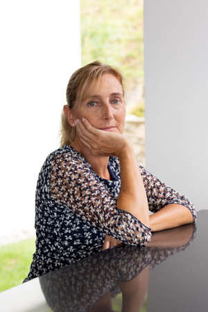 Portrait of a middle-aged woman with blond hair in a ponytail with a window with a green lawn and a white wall behind her. Reflective table and white backgroundの写真素材