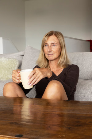 Attractive housewife in her fifties, pausing and relaxing in the living room with a cup of tea as she thinks about her past and future lifeの写真素材
