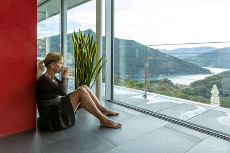 Beautiful young business woman pausing at her home, sitting on the ground while looking out the window in not too good weather but in the background you can see Lake Ceresio. We are in Switzerlandの素材 [FY310190894135]