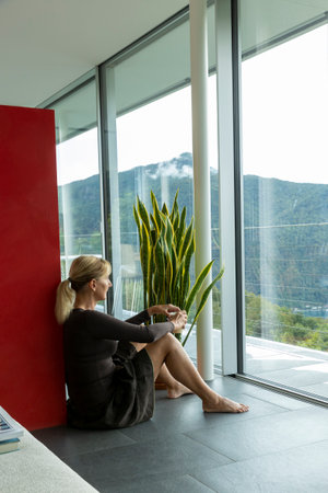Beautiful young business woman pausing at her home, sitting on the ground while looking out the window in not too good weather but in the background you can see Lake Ceresio. We are in Switzerlandの写真素材