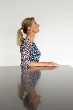 Portrait of a middle aged woman in profile with blonde hair tied in a ponytail and a blue and white polka dot shirt. She has her eyes open and is looking to the right. Reflective table and white backgroundの写真素材