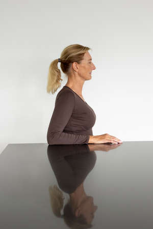 Middle aged woman portrait in profile with blonde hair tied in a ponytail with a white wall behind. She has her eyes closed and is looking to the right. Reflective table and white backgroundの写真素材