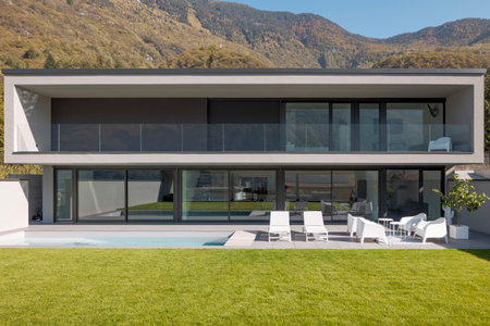 Front view modern house with two floors, pool two deck chairs and a small lounge with white armchairs. In front is a beautiful green lawn. Nobody insideの写真素材