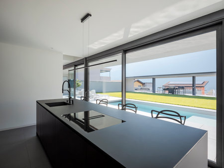 Black modern kitchen with sink and stools. There are bright windows where lots of light comes in and you can see the pool and garden. Nobody insideの写真素材