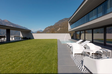 Modern house with manicured green garden, shady patio. Pool with two deck chairs and a small lounge with white armchairs. View of the Swiss Alps. Nobody insideの写真素材