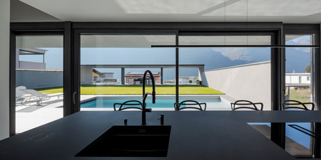 Black modern kitchen with sink and stools. There are bright windows where lots of light comes in and you can see the pool and garden. Nobody insideの写真素材
