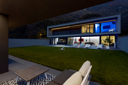 Modern house with swimming pool and garden in night scene illuminated by colored LED lights. In the foreground is a patio with a sofa. Nobody insideの写真素材