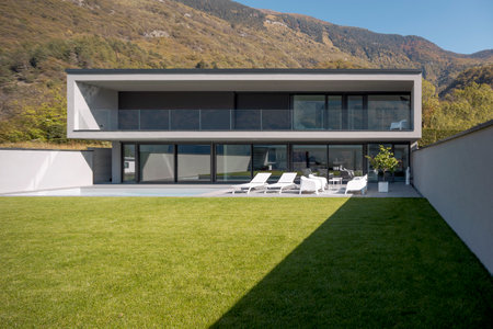 Front view modern house with two floors, pool two deck chairs and a small lounge with white armchairs. In front is a beautiful green lawn. Nobody insideの写真素材