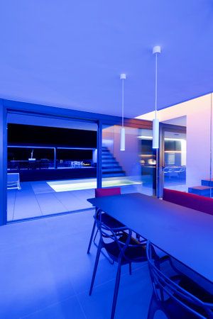 Liiving room with table and a red sofa lit by blue LEDs and lamps attached to the ceiling. Large window overlooking the lighted pool and garden. Nobody insideの写真素材