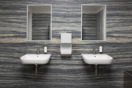 Two white sinks of an Italian public bathroom. The walls are gray white, one shaded. The photographic shot is frontal and the liquid soap holders are white.の写真素材