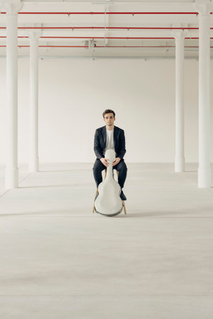 A young boy is sitting on a stool with his guitar case in front of him. Interior of a renovated former factory.の写真素材